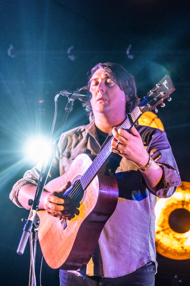 Een muzikant speelt gitaar tijdens een optreden met een fel verlichte achtergrond.