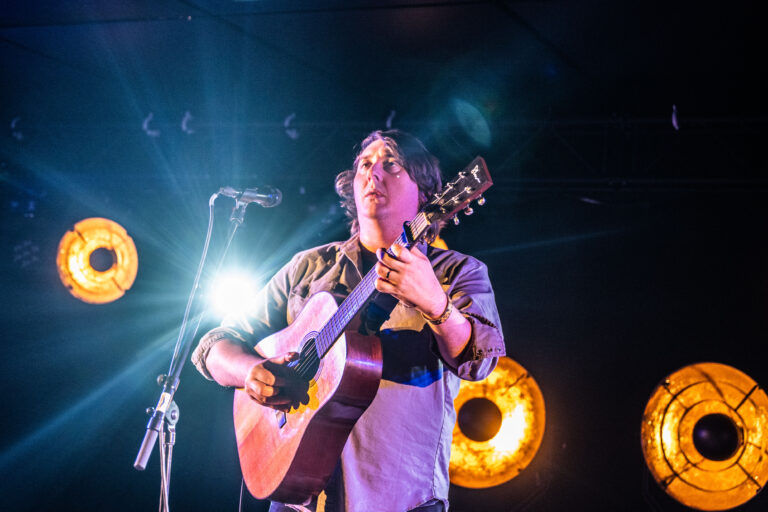 Een muzikant voert een nummer uit met een gitaar op het podium, omringd door fel licht.