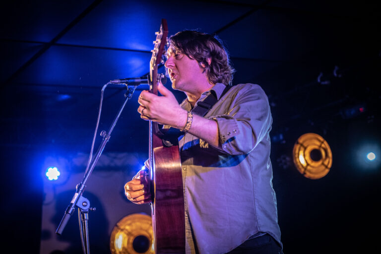Een muzikant speelt gitaar op een podium, omringd door blauwe en gouden lichten.