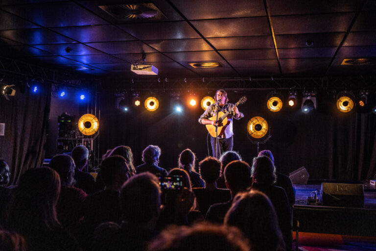 Een muzikant speelt gitaar op een podium terwijl een publiek naar hem kijkt, verlicht door warme lichten.