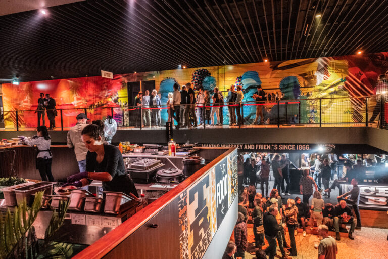 Een drukke bar met een bovenverdieping, waar gasten staan en een kleurrijke muurschildering op de achtergrond zichtbaar is.