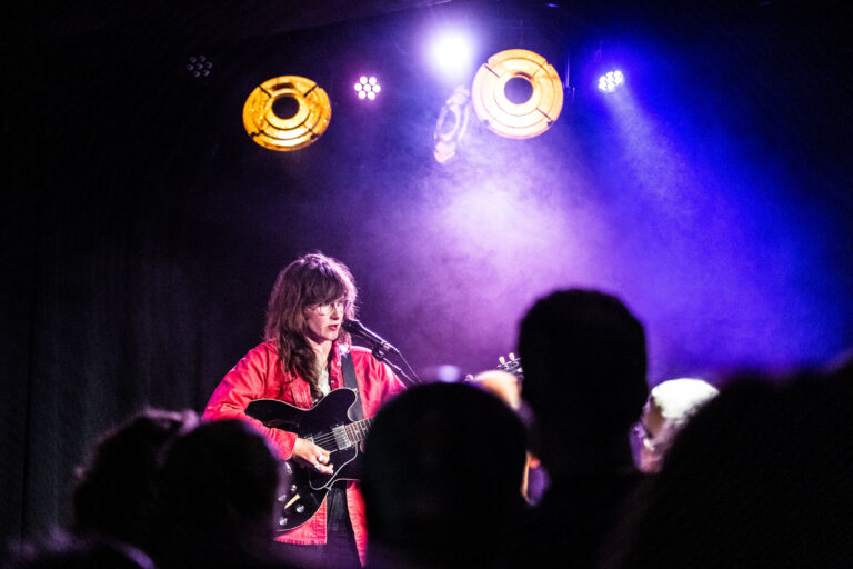 Een muzikante in een rode jas speelt gitaar en zingt op een podium met kleurrijke verlichting.