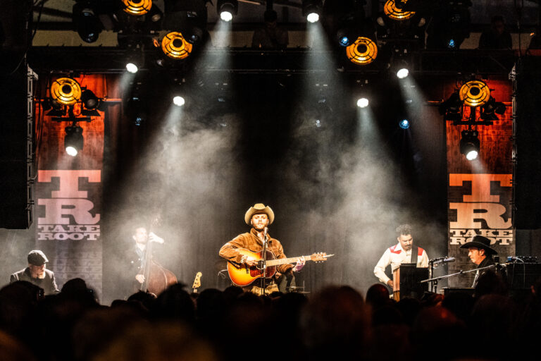 Een muzikant speelt gitaar op een podium met spotlights en een rookachtige atmosfeer.