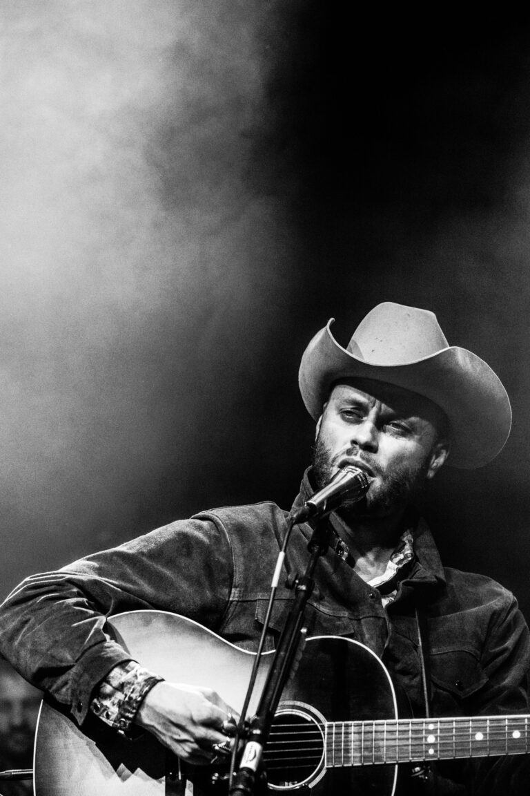 Zwart-wit afbeelding van een man in een cowboyhoed die een gitaar speelt en zingt op een podium.