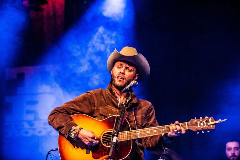 Een muzikant speelt gitaar en zingt op een podium met blauwe verlichting.