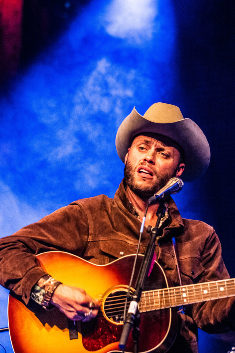 Een muzikant met een hoed speelt gitaar en zingt op een podium met een kleurrijke achtergrond.