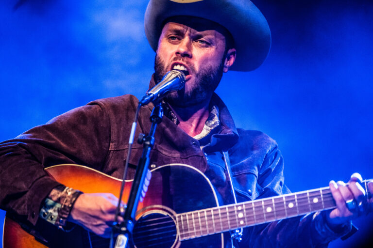 Een man met een hoed speelt gitaar en zingt op een podium, omgeven door blauwe verlichting.