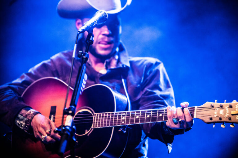 Een muzikant speelt gitaar op een podium, gekleed in een hoed en een donkere outfit, met een rokerige achtergrond.