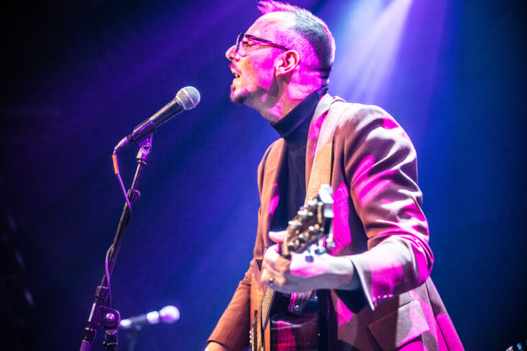 Een muzikant met een gitaar zingt in een fel verlichte concertzaal.