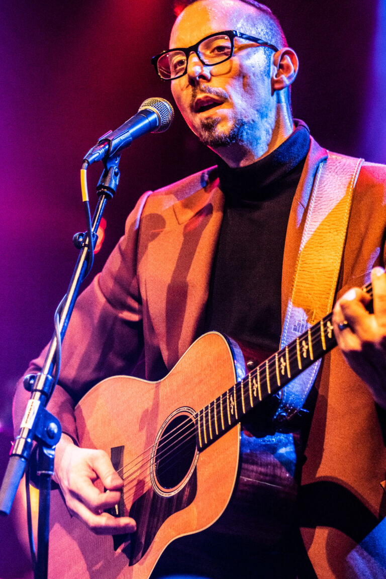 Een muzikant zingt en speelt gitaar op een podium met kleurrijke verlichting.