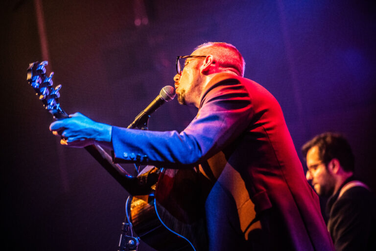Een muzikant met een gitaar zingt in een sfeer van kleurrijke verlichting op het podium.