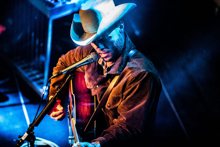 Een muzikant in een cowboyhoed speelt gitaar op het podium met theatrale verlichting.