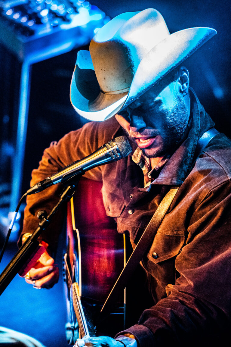 Een muzikant met een cowboyhoed speelt gitaar en zingt in een sfeervolle omgeving.