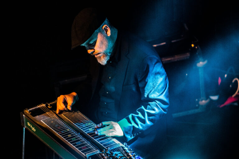 Een muzikant speelt op een pedal steel gitaar, omgeven door een sfeervolle, blauwe belichting.