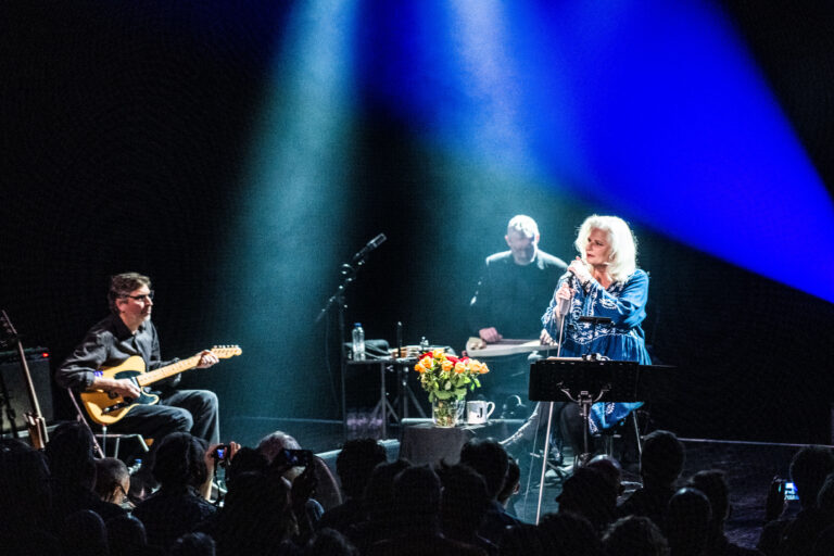Een muzikale uitvoering met drie artiesten op het podium, verlicht door een blauwe spotlight, met bloemen op een tafel.