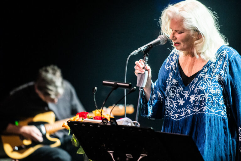 Een vrouw met blonde, krullende haren zingt in een microfoon, terwijl een muzikant op de achtergrond gitaar speelt.
