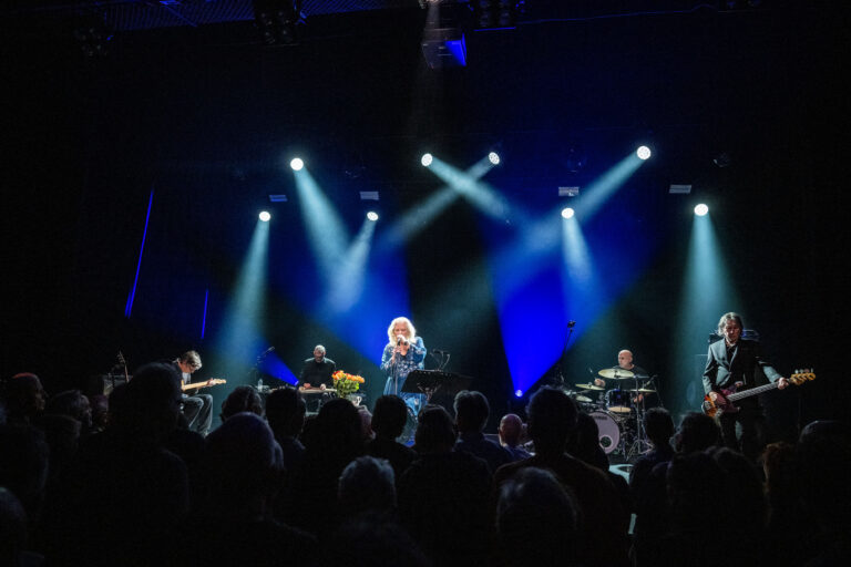 Een muzikant staat op een podium met bandleden, omgeven door een menigte en sfeervolle verlichting.