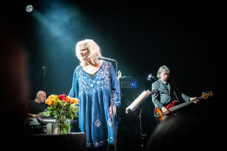 Een vrouw in een blauwe jurk staat op een podium met een microfoon, terwijl een muzikant op de achtergrond een gitaar bespeelt, vergezeld door een boeket bloemen.