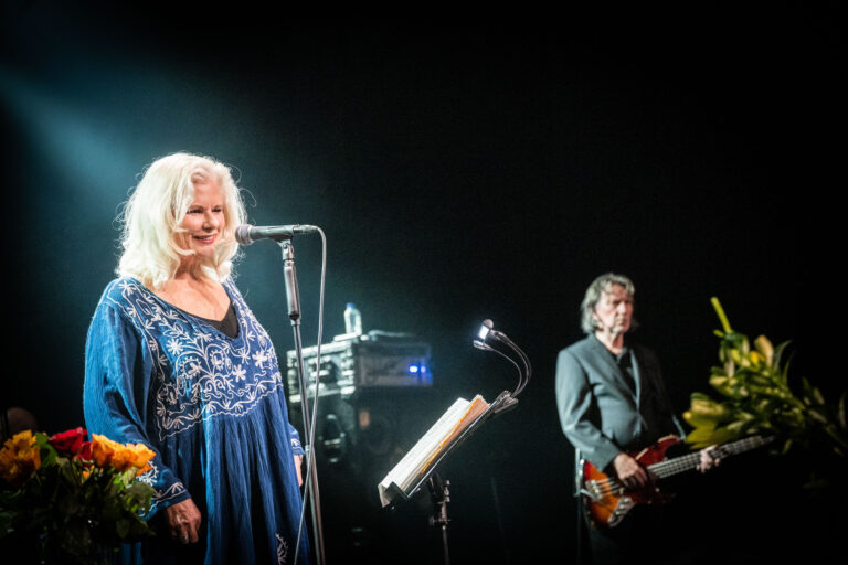 Een zangeres met een blauwe jurk staat op het podium, terwijl een muzikant met een gitaar achter haar speelt.