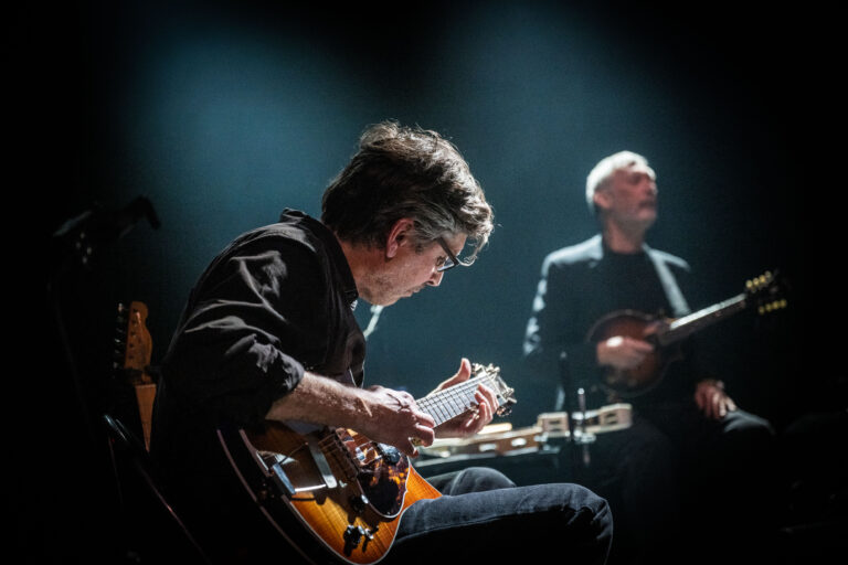 Een muzikant speelt gitaar op een podium, terwijl een andere artiest erbij staat.