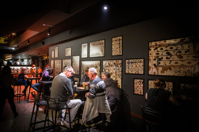 Een groep mensen zit aan hoge tafels in een bar met kunstwerken aan de muur.