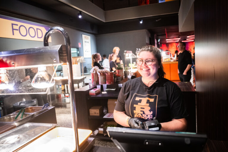 Een werknemer staat achter een toonbank in een restaurant, glimlachend naar de camera, terwijl anderen in de achtergrond bezig zijn.