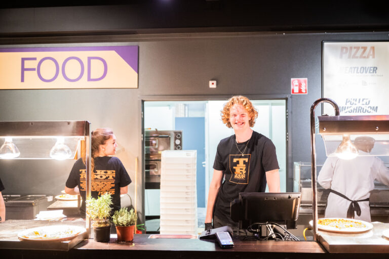 Twee medewerkers staan achter de balie van een eetstand, met een focus op hun vrolijke uitdrukkingen.