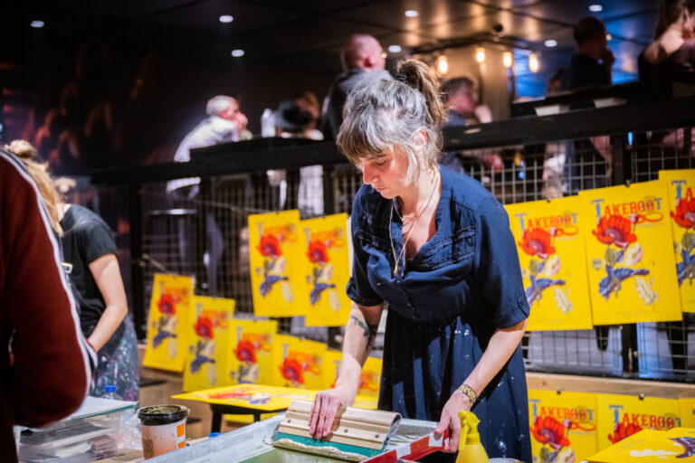 Een vrouw in een blauwe jurk die printtechnieken toepast, met kleurrijke kunstwerken op de achtergrond.