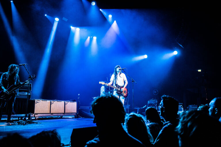 Een muzikant staat op het podium met een gitaar, omringd door blauw licht en een menigte kijkers.