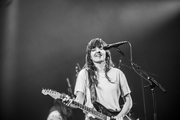 Een artiest op het podium met een elektrische gitaar en een microfoon, lachend tijdens een optreden.