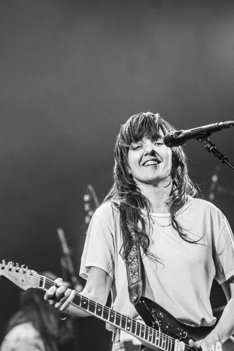 Zwarte en witte afbeelding van een muzikante die lacht en in de microfoon zingt terwijl ze een gitaar vasthoudt.