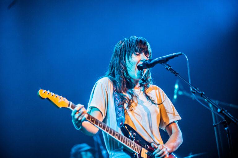 Een muzikant zingt en speelt gitaar op een podium met een blauwe achtergrondverlichting.