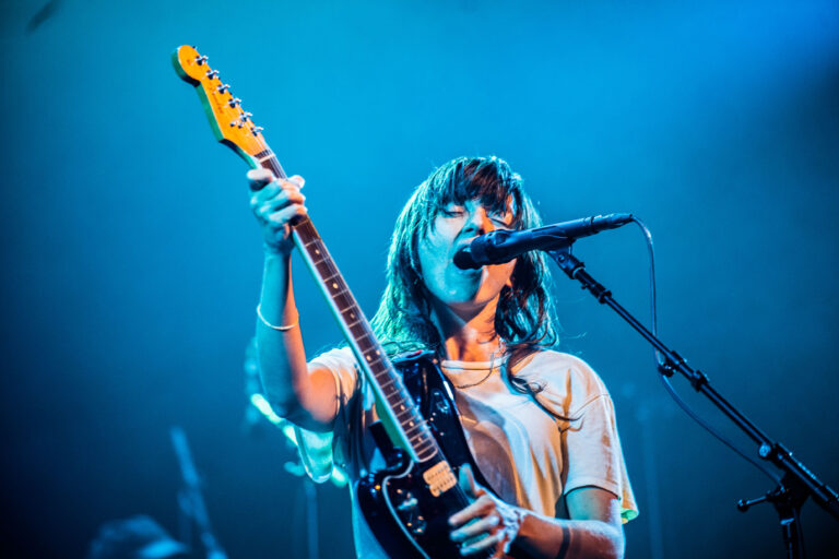 Een muzikant met een gitaar zingt in een levendig blauw verlichte sfeer op het podium.