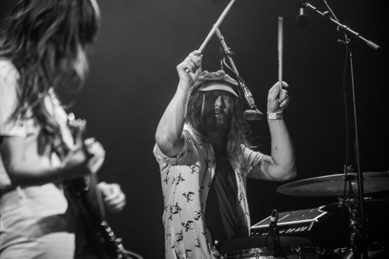 Een drummer met lang haar speelt enthousiast op een drumstel tijdens een optreden.