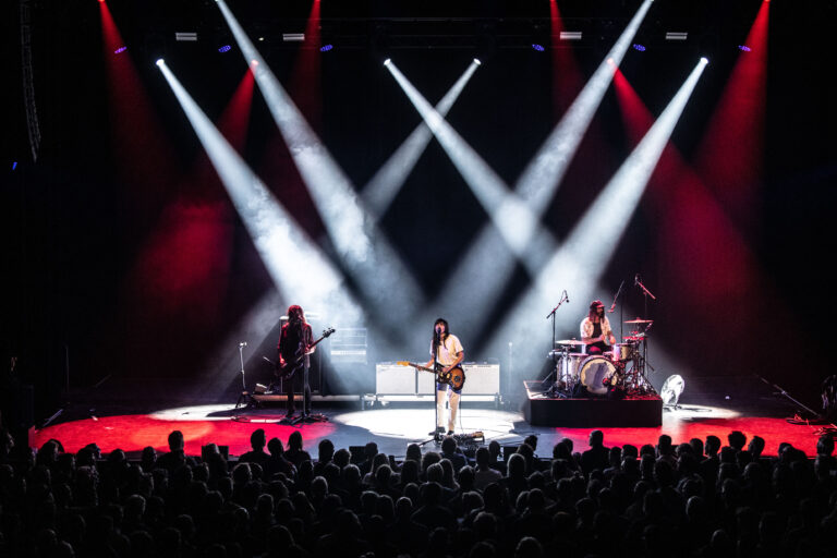 Een muzikale optreden met een band op het podium, omringd door krachtige lichtstralen en een publiek dat geniet.