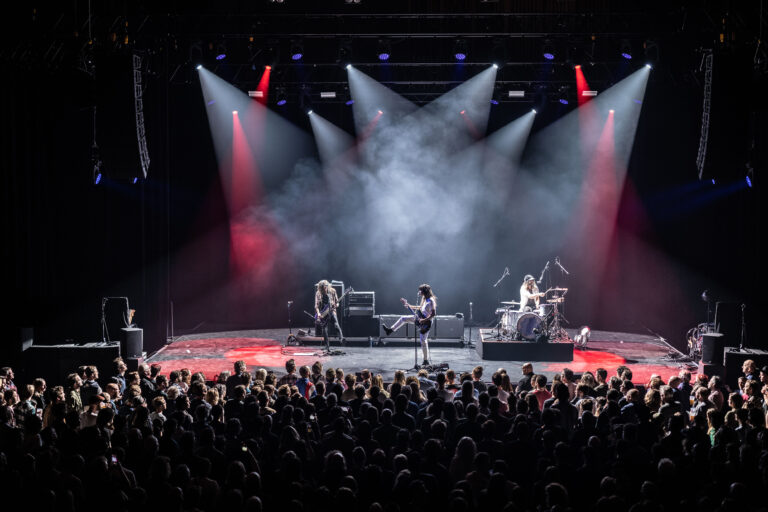 Een live band optredend op een podium voor een menigte met kleurrijke lichten en rook.