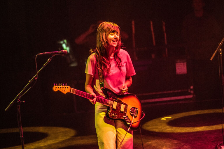 Een muzikante met een elektrische gitaar staat lachend op het podium, omringd door sfeervolle verlichting.