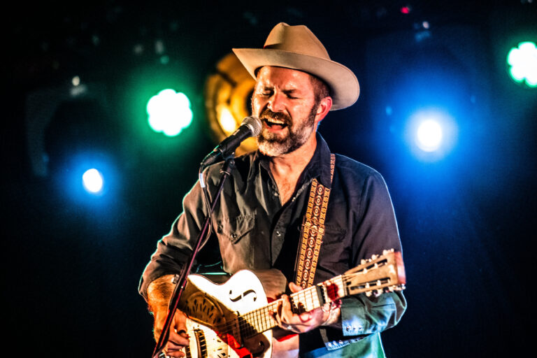 Een muzikant met een hoed speelt gitaar op een podium, omringd door kleurrijke lichten.