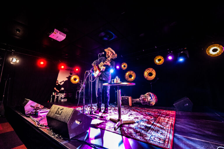 Een muzikant speelt gitaar op een podium met kleurrijke verlichting en een sfeervolle achtergrond.