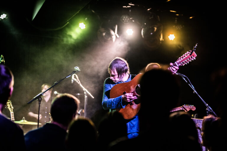 Een muzikant speelt op het podium terwijl het publiek toekijkt en er kleurrijke lichten van boven hangen.