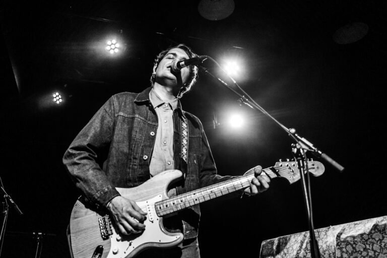 Een muzikant speelt gitaar en zingt op een podium met lichteffecten.