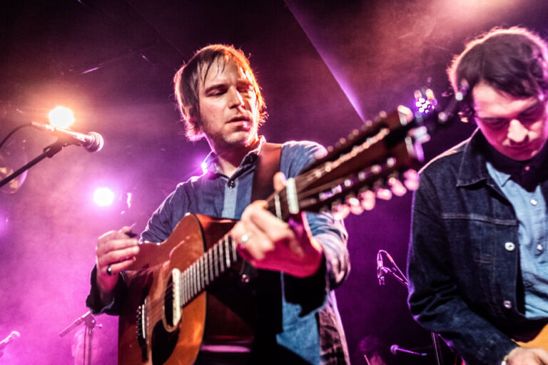 Een muzikant speelt gitaar op het podium met kleurrijke lichten op de achtergrond.