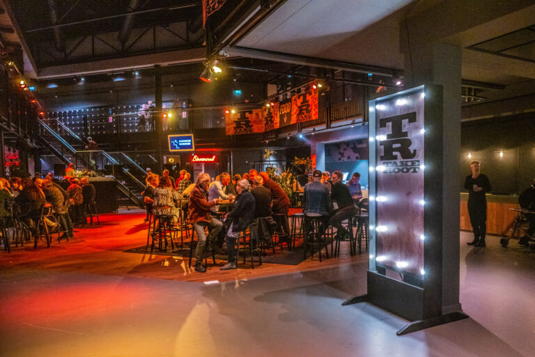 Een drukke ruimte met sfeervolle verlichting en een groep mensen die aan hoge tafels zit.