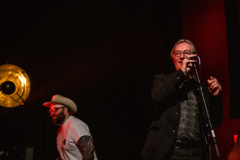 Twee muzikanten treden op op een podium, met een focus op de zanger aan de rechterkant die een microfoon vasthoudt.
