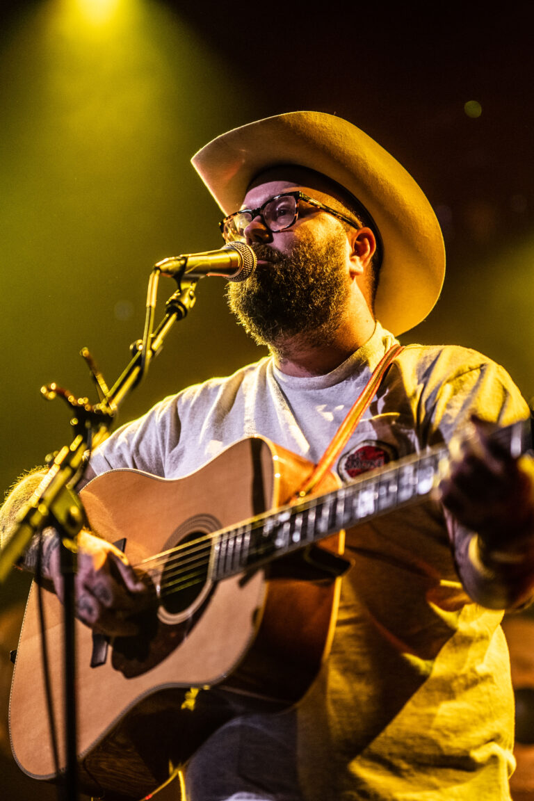 Een muzikant met een hoed speelt gitaar en zingt in een sfeervolle setting met gele verlichting.