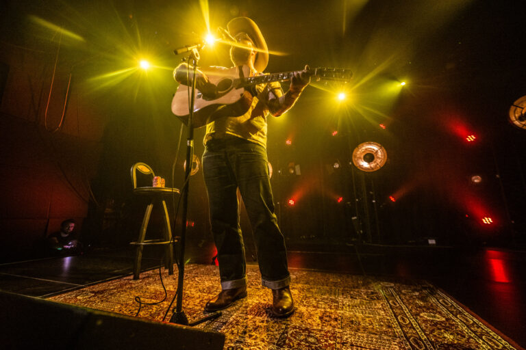 Een muzikant staat op het podium en speelt gitaar, omringd door sfeervolle gele verlichting.