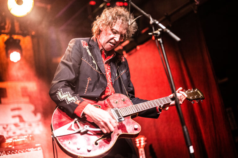 Een muzikant speelt live op het podium met een rode gitaar en heeft een expressieve uitstraling.