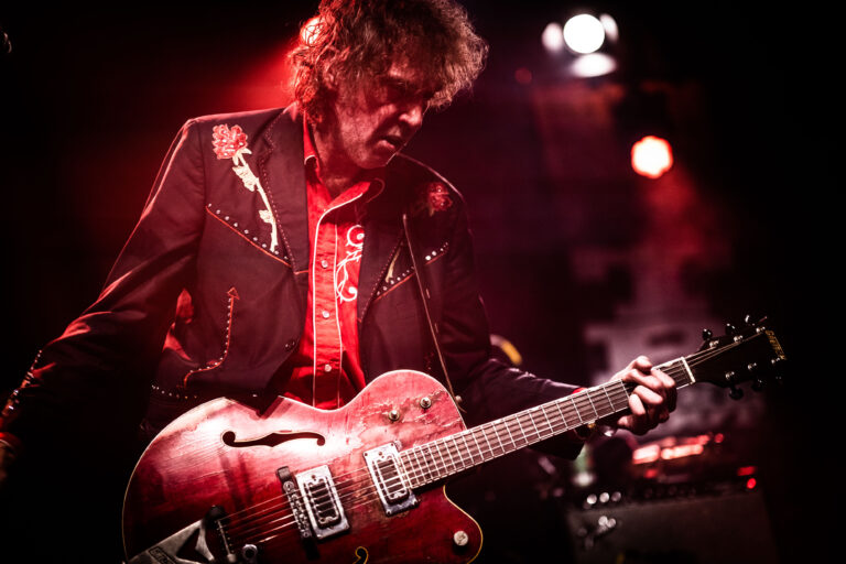 Een muzikant speelt op een rode gitaar tijdens een optreden met sfeerverlichting.