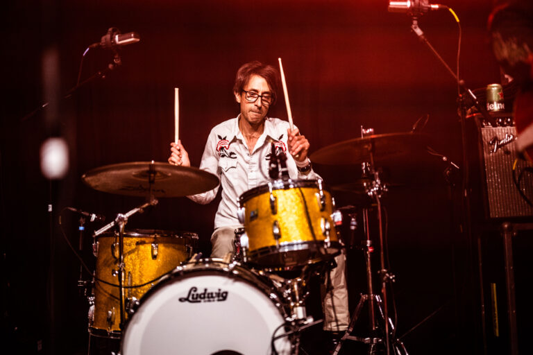 Een drummer speelt op een set gele en witte drums in een sfeervolle, verlichte omgeving.