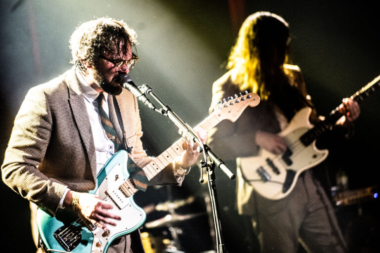 Twee muzikanten spelen op een podium, één zingt en bespeelt een blauwe gitaar, terwijl de ander een lichte basgitaar vasthoudt, omringd door sfeervolle belichting.